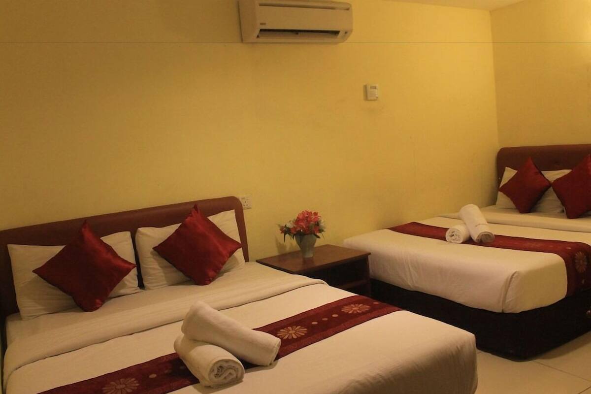 A hotel room with two beds, each with red pillows and white bedding, a small table with a vase of flowers, and an air conditioning unit on the wall.