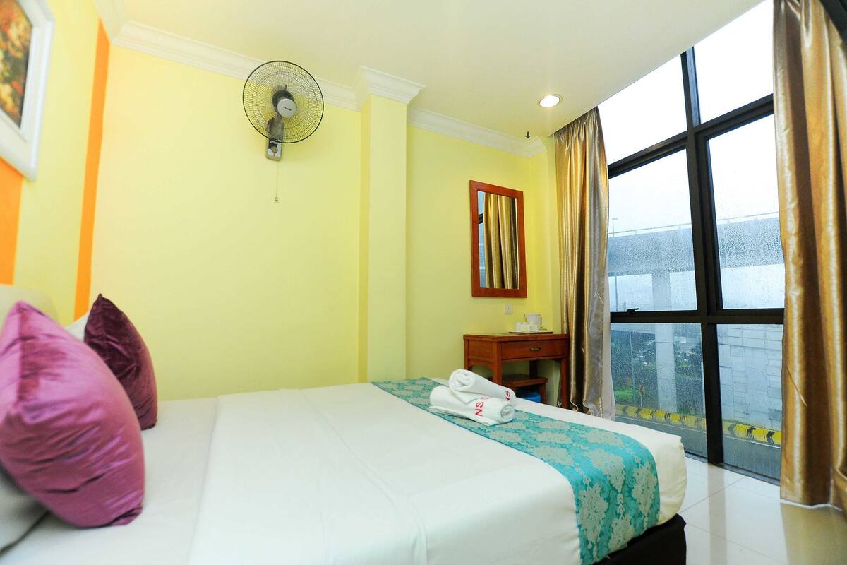A hotel room with a bed, pillows, a bedside table, a fan, a window with curtains, and a view of the outside.