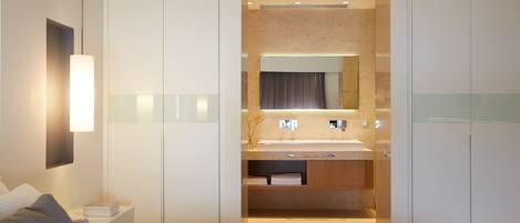 A modern hotel room with a large bed, a bedside table, a nightstand, a built-in vanity with a mirror, and a shower area.