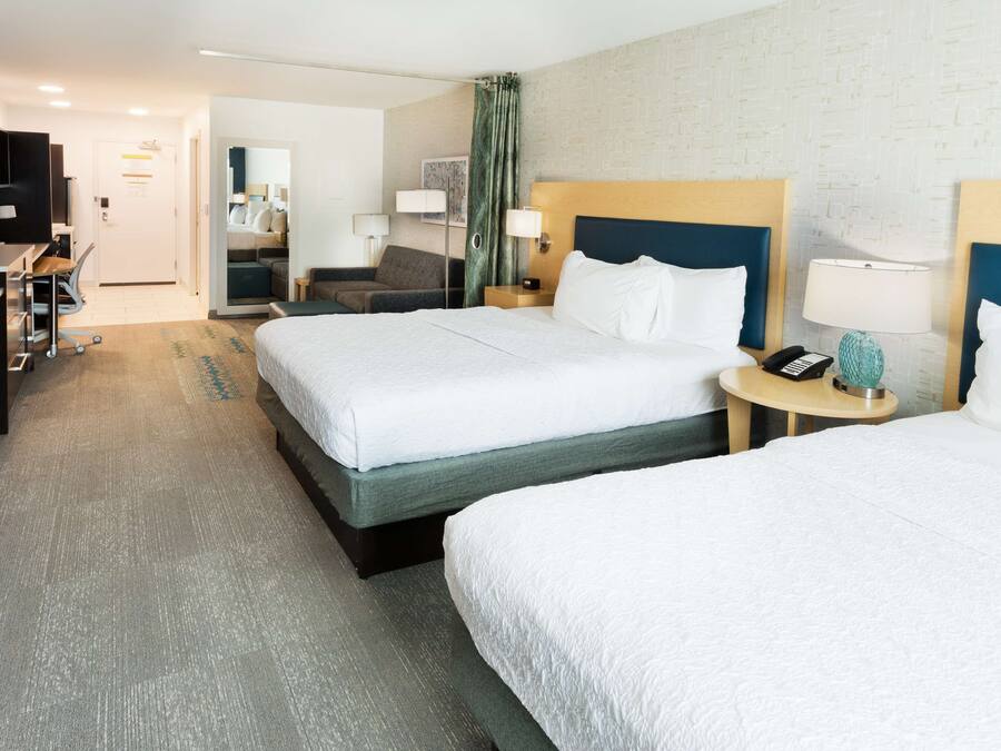 A hotel room with two beds, a desk, a chair, a TV, and a small table with a lamp.