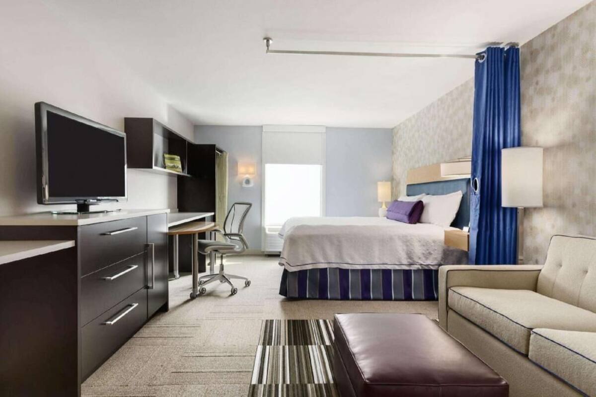 A hotel room with a bed, a desk with a computer, a TV, a sofa, and a window.