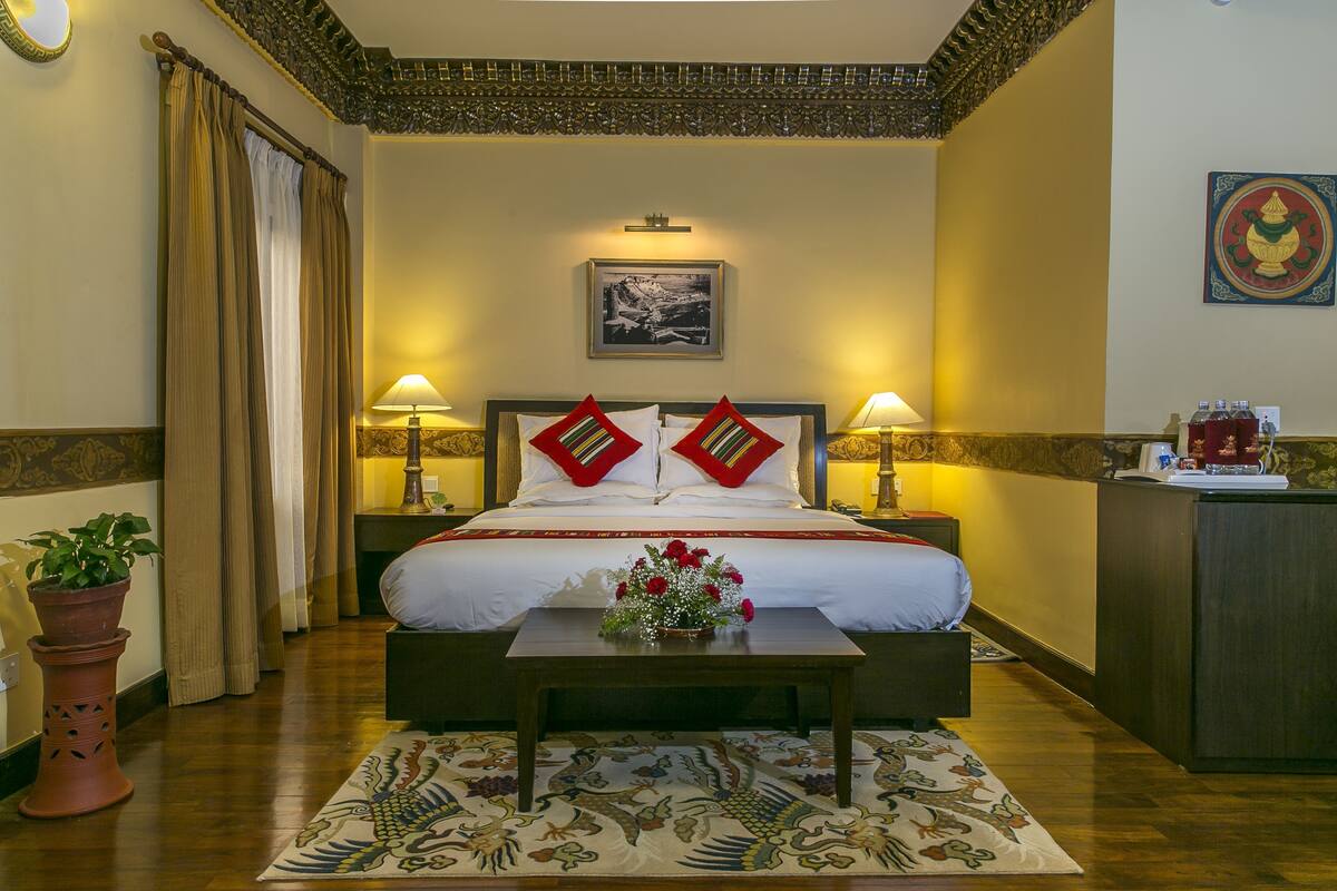 A hotel room with a bed, bedside lamps, a desk, a potted plant, and a decorative rug.