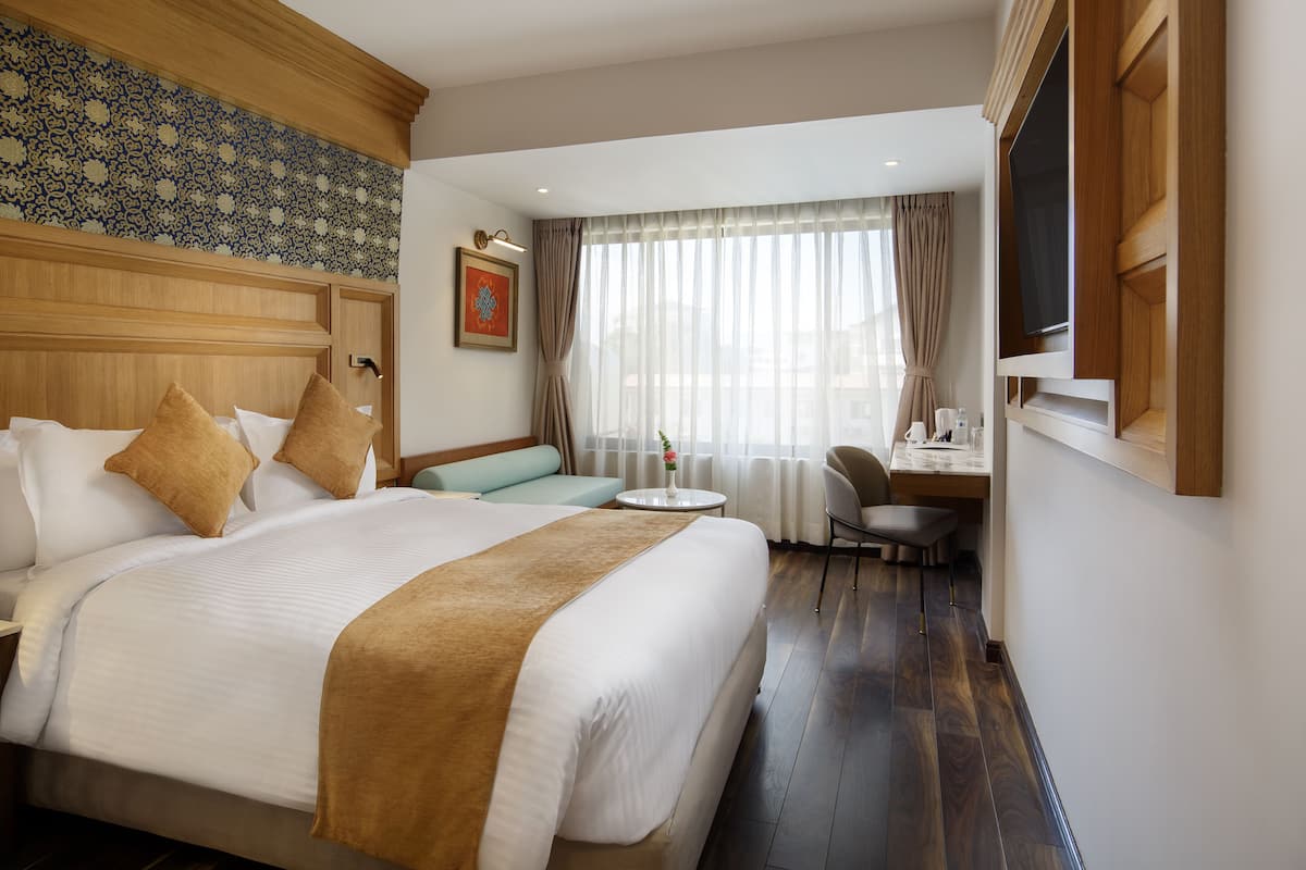 A hotel room with a large bed, a desk, a chair, a small round table, and a window with curtains.