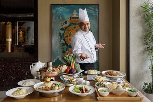 A chef standing in front of a table with various dishes and a traditional painting in the background.