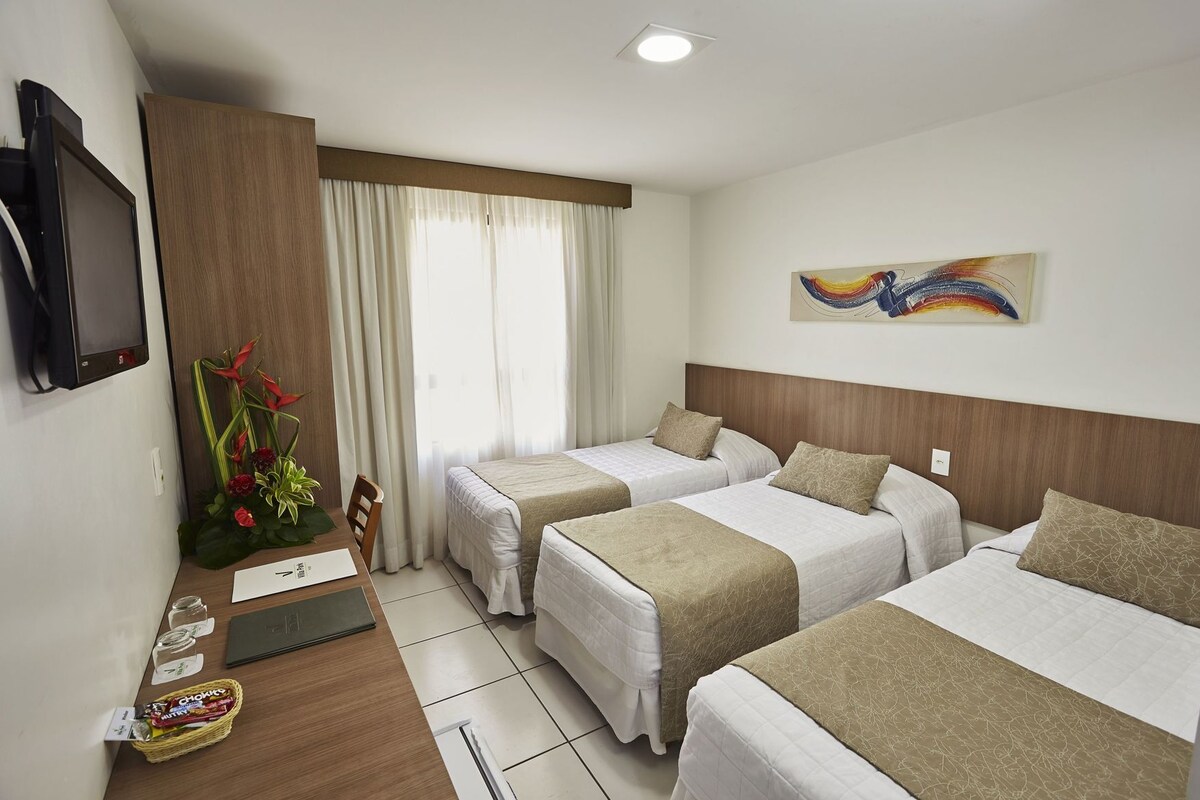 Habitación de hotel con dos camas, televisión, escritorio con computadora, ventana con cortinas y un cuadro en la pared.