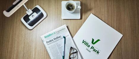 A wooden table with a white folder labeled "Villa Park Hotel," a newspaper with a pen and glasses, a smartphone, and a cup of coffee.