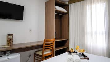 A hotel room with a bed, a wooden wardrobe, a desk with a chair, a television mounted on the wall, and a window with curtains.