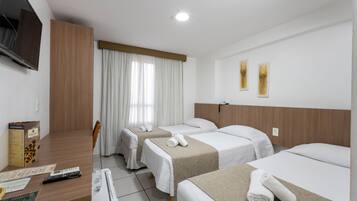 Quarto de hotel com três camas, uma televisão na parede, uma secretária com computador e controles remotos, e uma janela com cortinas.
