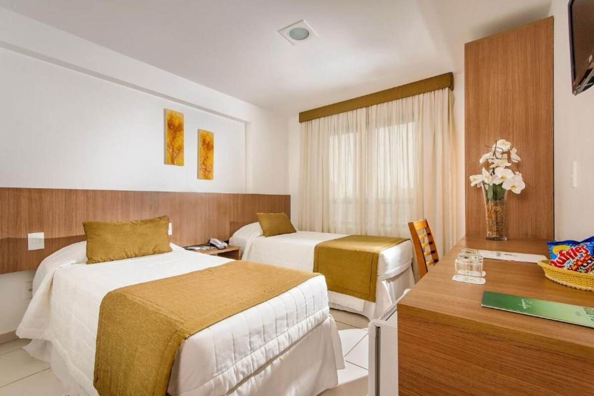 Une chambre d’hôtel avec deux lits, une tête de lit en bois, un bureau avec une chaise, un vase avec des fleurs et un panier de collations.