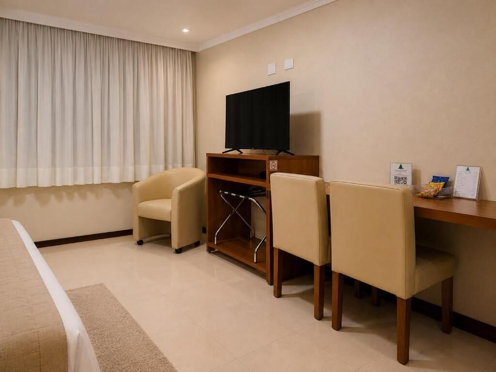 A hotel room with a bed, a desk with a computer, two chairs, a television, and a window with curtains.