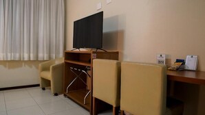 A hotel room with a television on a wooden stand, a desk with a chair, a small table with a sign, and a curtain. - Cedro Hotel (Londrina)