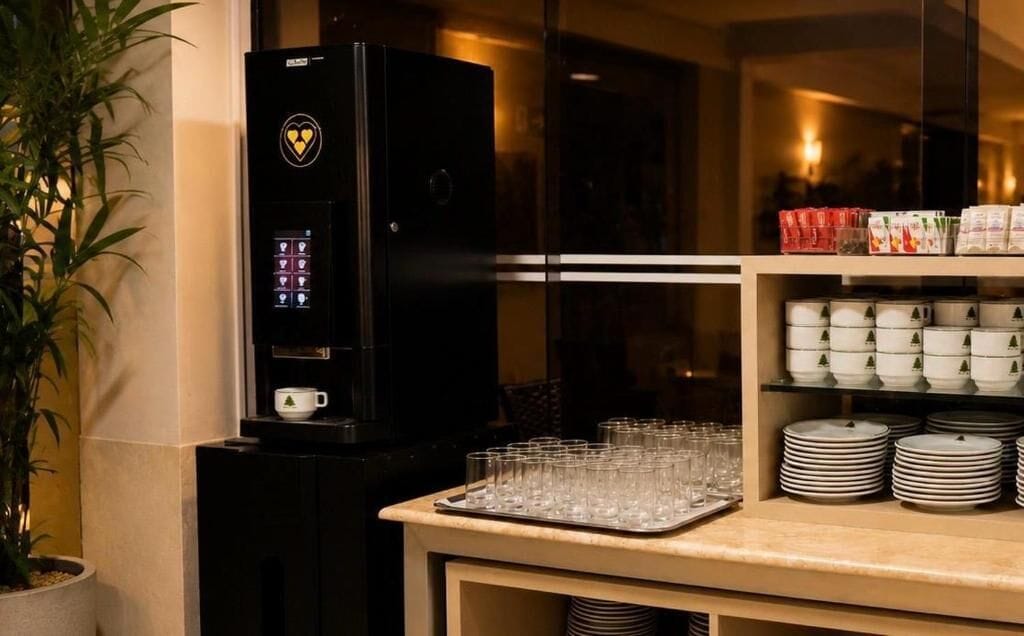 A coffee machine with a digital interface, a tray of glasses, and a shelf with stacked plates and cups.