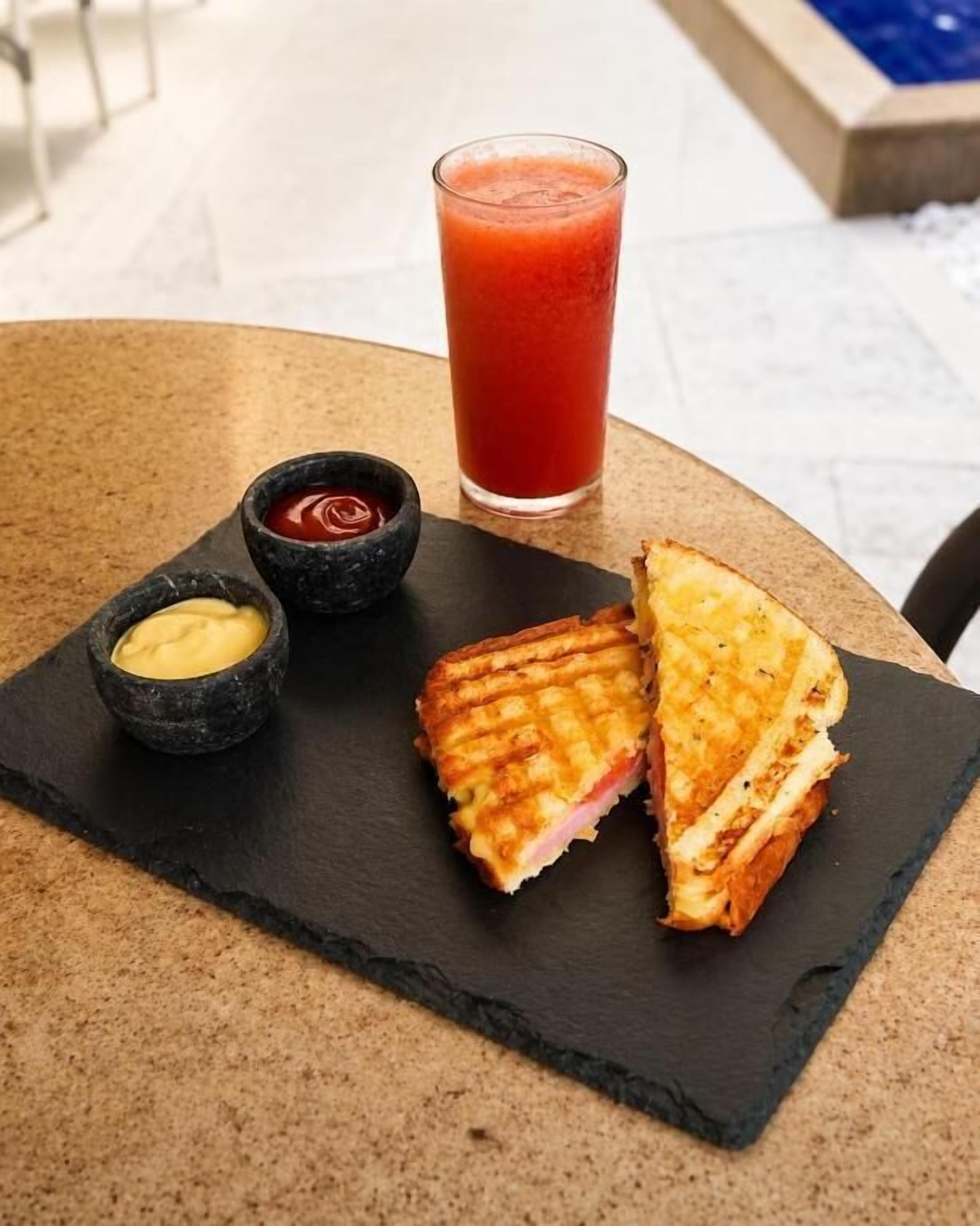 A sandwich on a slate board with a glass of red drink and two small bowls of condiments.