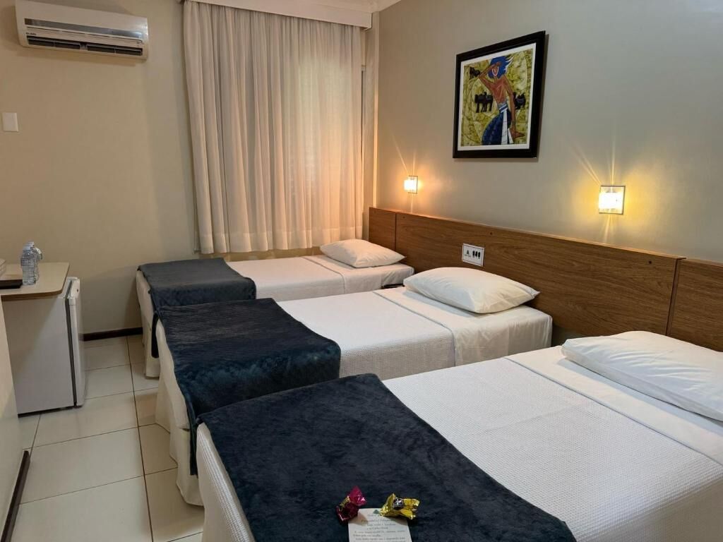 A hotel room with three beds, a desk, an air conditioner, and a painting on the wall.