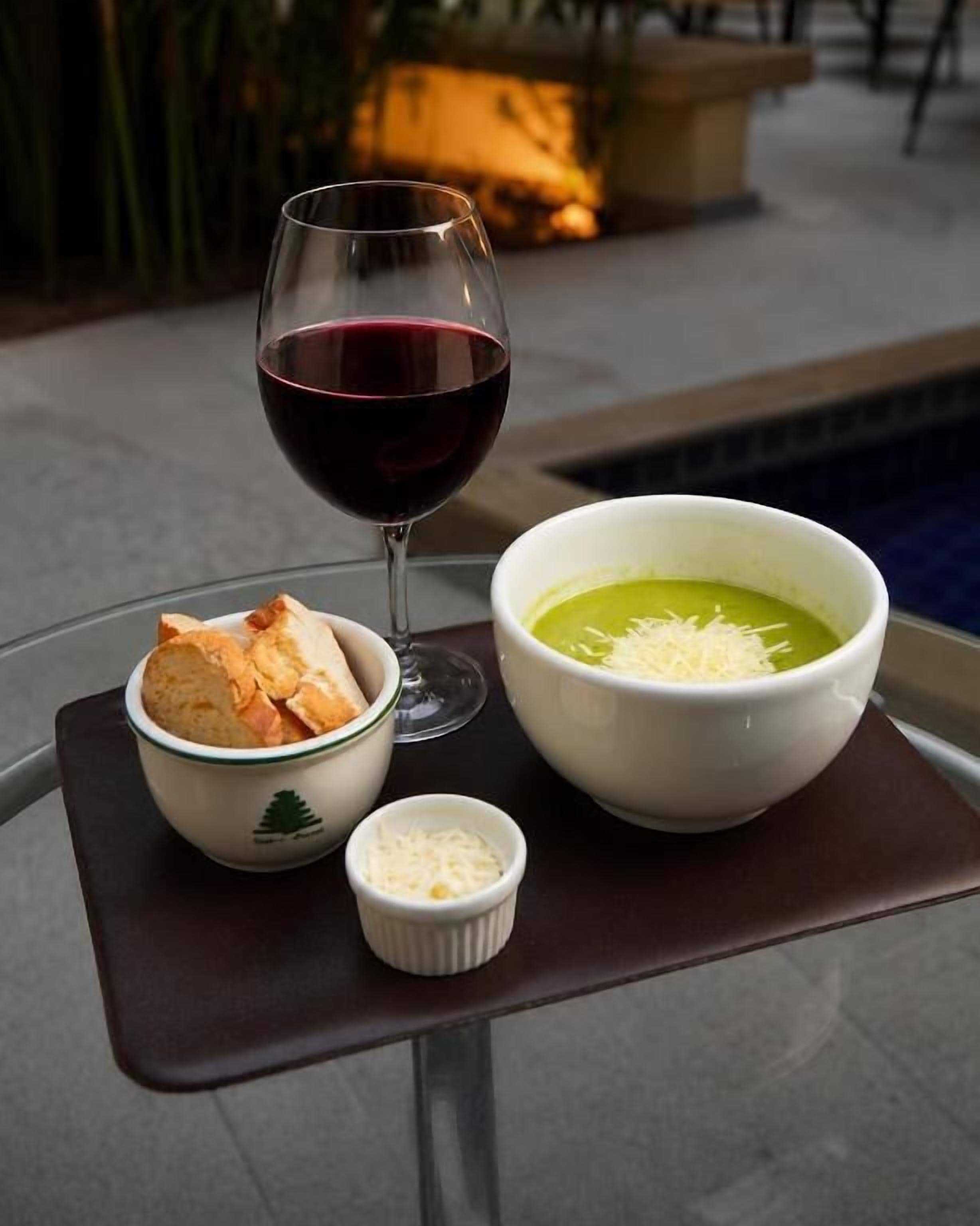 A dining table with a glass of red wine, a bowl of soup, a bowl of cheese, and a small dish of bread.