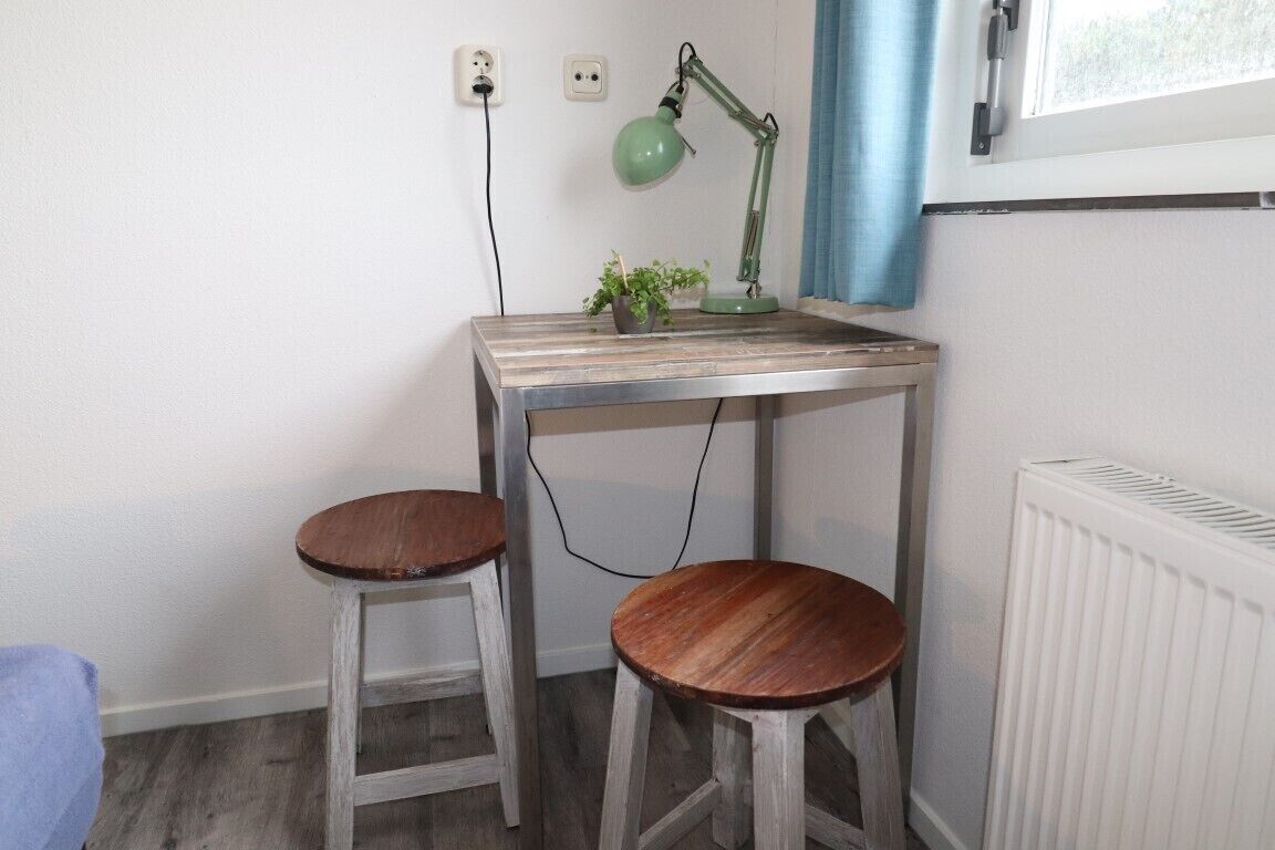 A small workspace with a desk and two stools, a green desk lamp, a potted plant, and a window with blue curtains.