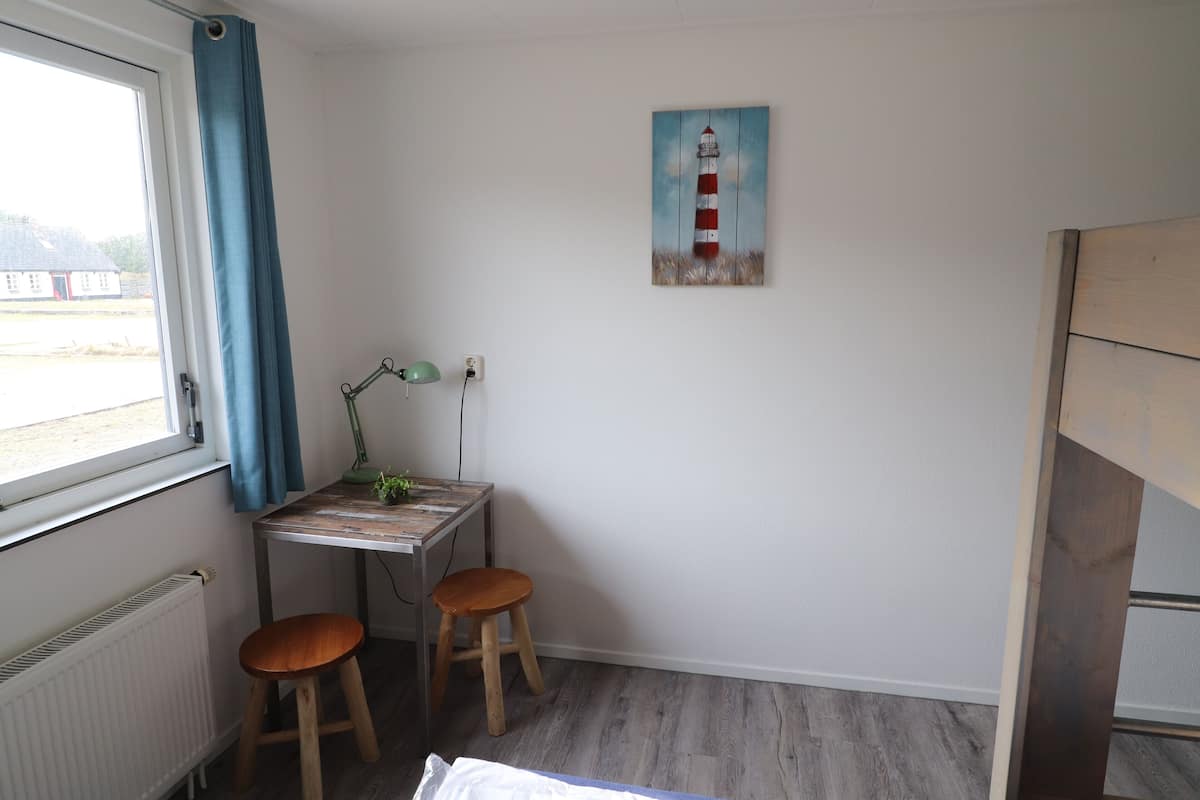 A small room with a desk, two stools, a radiator, a window with curtains, and a painting of a lighthouse.