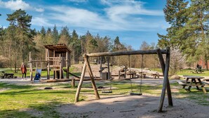 Une structure de jeux en bois rustique, dotée de balançoires et d’un toboggan, entourée de tables et de bancs de pique-nique, le tout situé dans un parc.