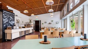 Un grand espace dédié à la salle à manger, avec des tables vert clair et des chaises en bois, un tableau noir où sont écrits des mots liés à la nourriture, et un plafond moderne agrémenté de luminaires suspendus.