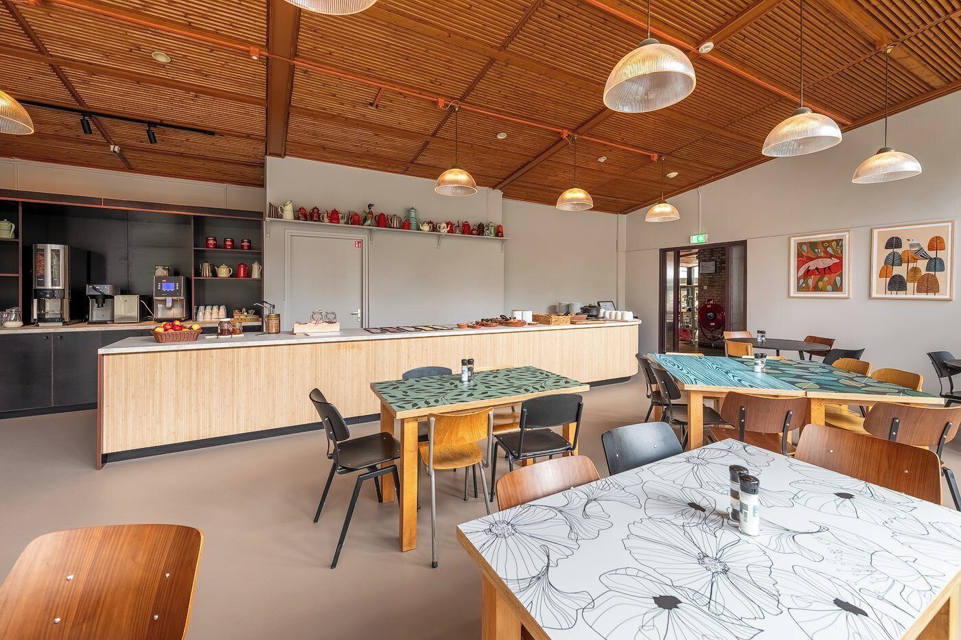 Ein modernes Café mit Holzbalkendecke, einer zentralen Theke mit verschiedenen Angeboten und Tischen mit blumigen Tischdecken.