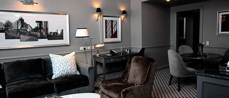 Una habitación de hotel moderna con suelo a cuadros blanco y negro, un sofá negro con cojín estampado, un sillón marrón, una mesa redonda blanca y un escritorio con lámpara.