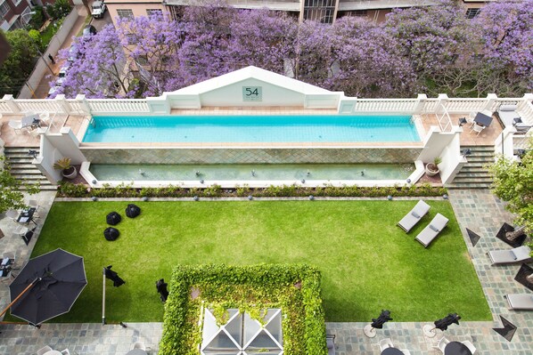 Una piscina en la azotea con un letrero con el número '54', rodeada de un césped cuidado, asientos al aire libre y vistas a árboles con flores moradas.