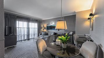 A modern hotel room with a dining area, a flat-screen TV, a sofa, a coffee table, and a potted plant.
