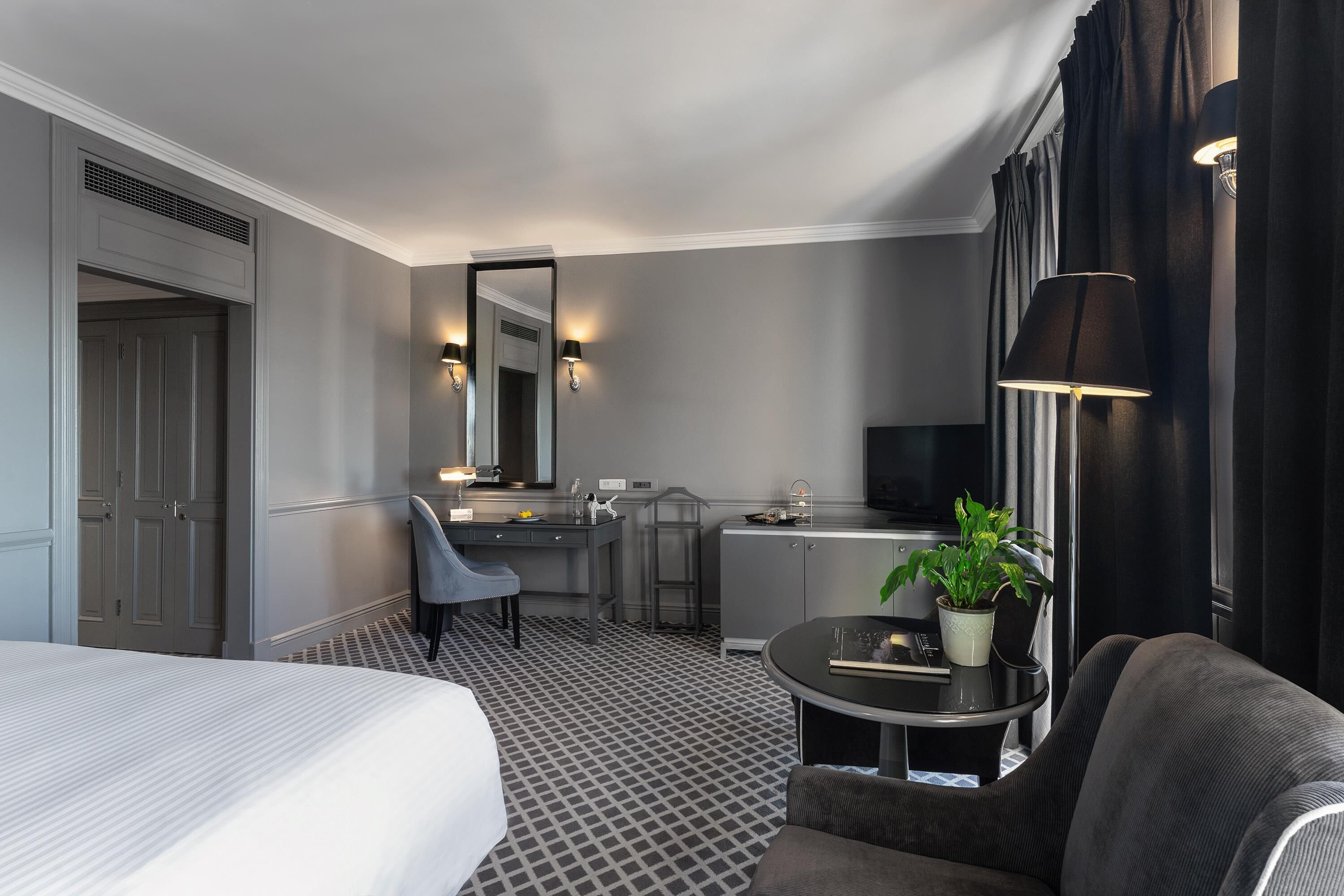 A hotel room with a bed, a desk, a chair, a small table with a plant, and a television.