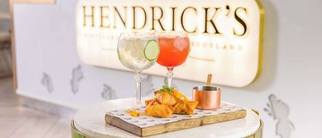 A table with two glasses of beverages, a small copper pot, and a plate of food on a round table with a gold base, set against a wall with a sign reading "HENDRICK'S".
