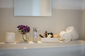 A bathroom with a white sink, a vase with purple flowers, a mirror, and rolled towels. - Santacroce Luxury Rooms (Lecce)