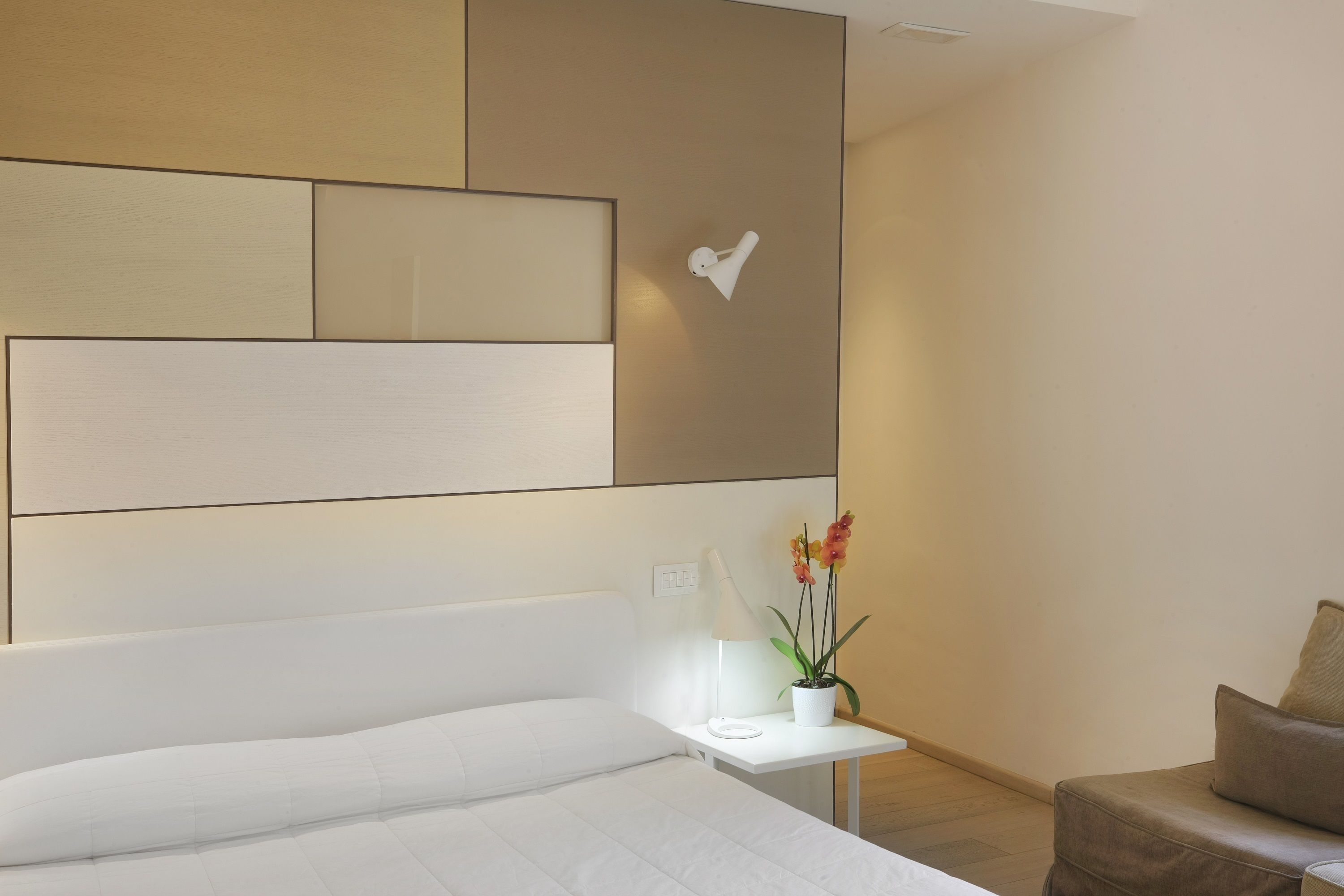Una camera d'hotel moderna con un letto grande, un comodino con una lampada, una pianta in vaso e un punto luce a parete.