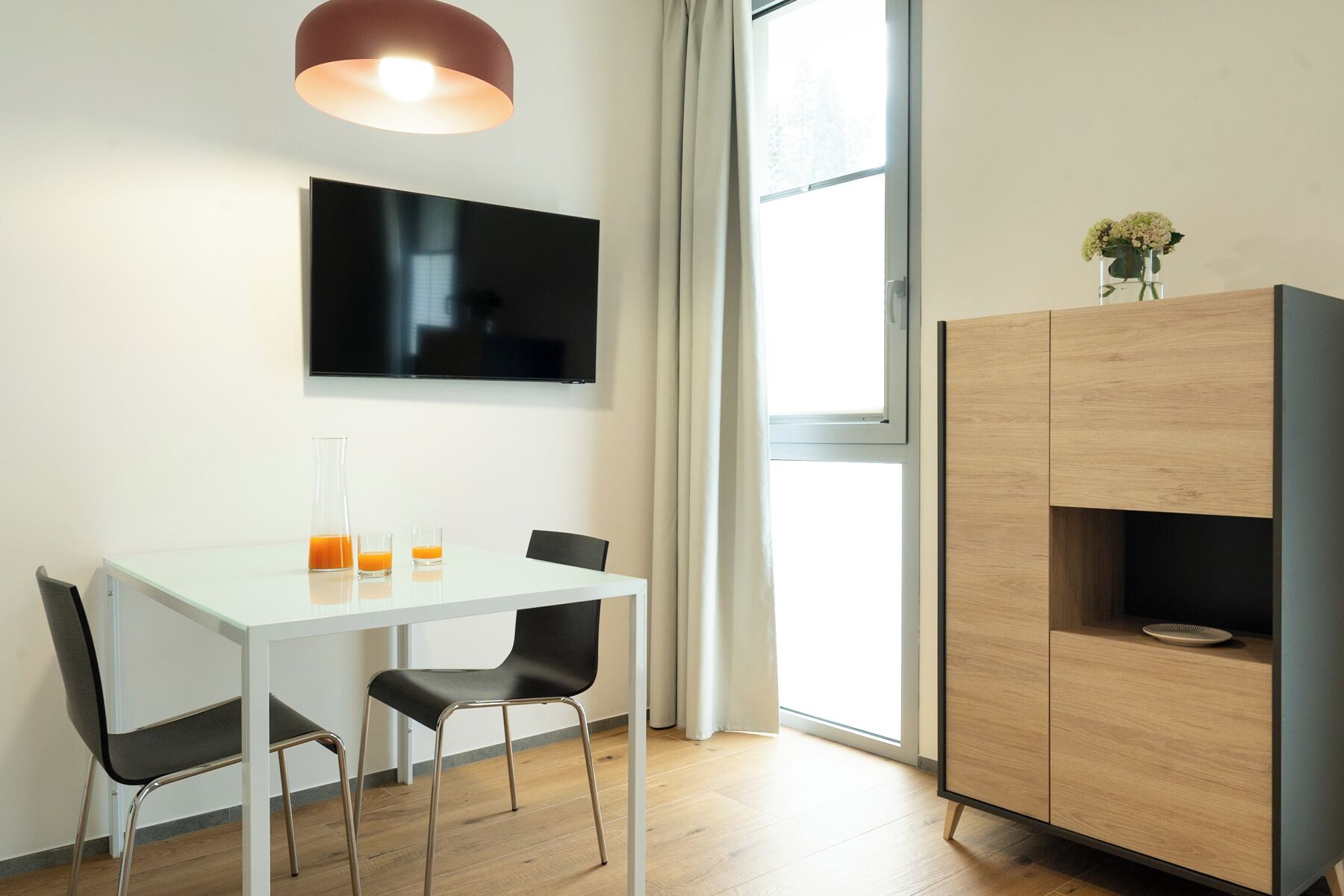 A modern dining area with a white table, two black chairs, a flat-screen TV, a wooden cabinet, and a large window with curtains.