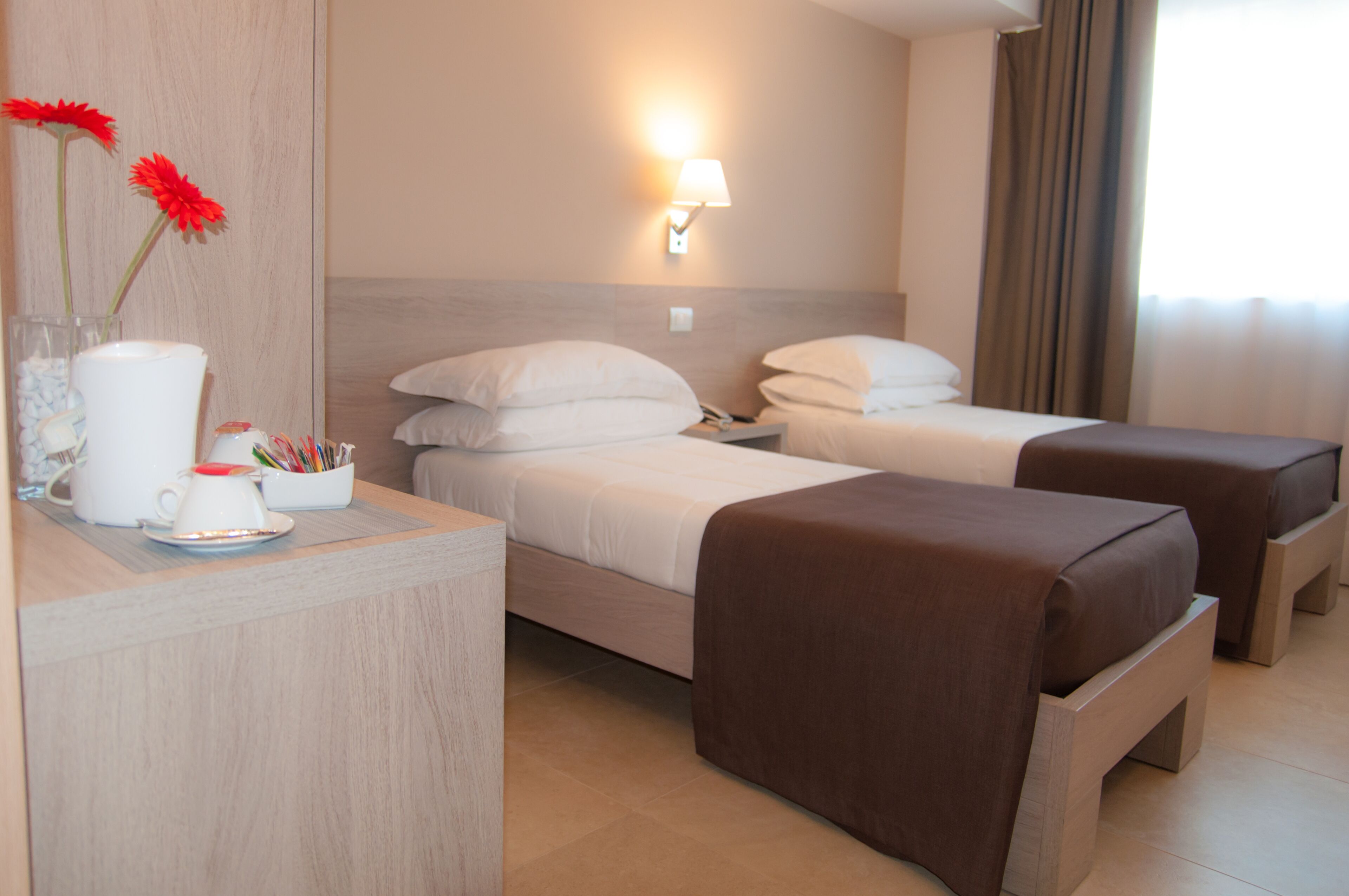 Una habitación de hotel con dos camas, una mesita de noche con lámpara, un florero con flores y una cafetera.