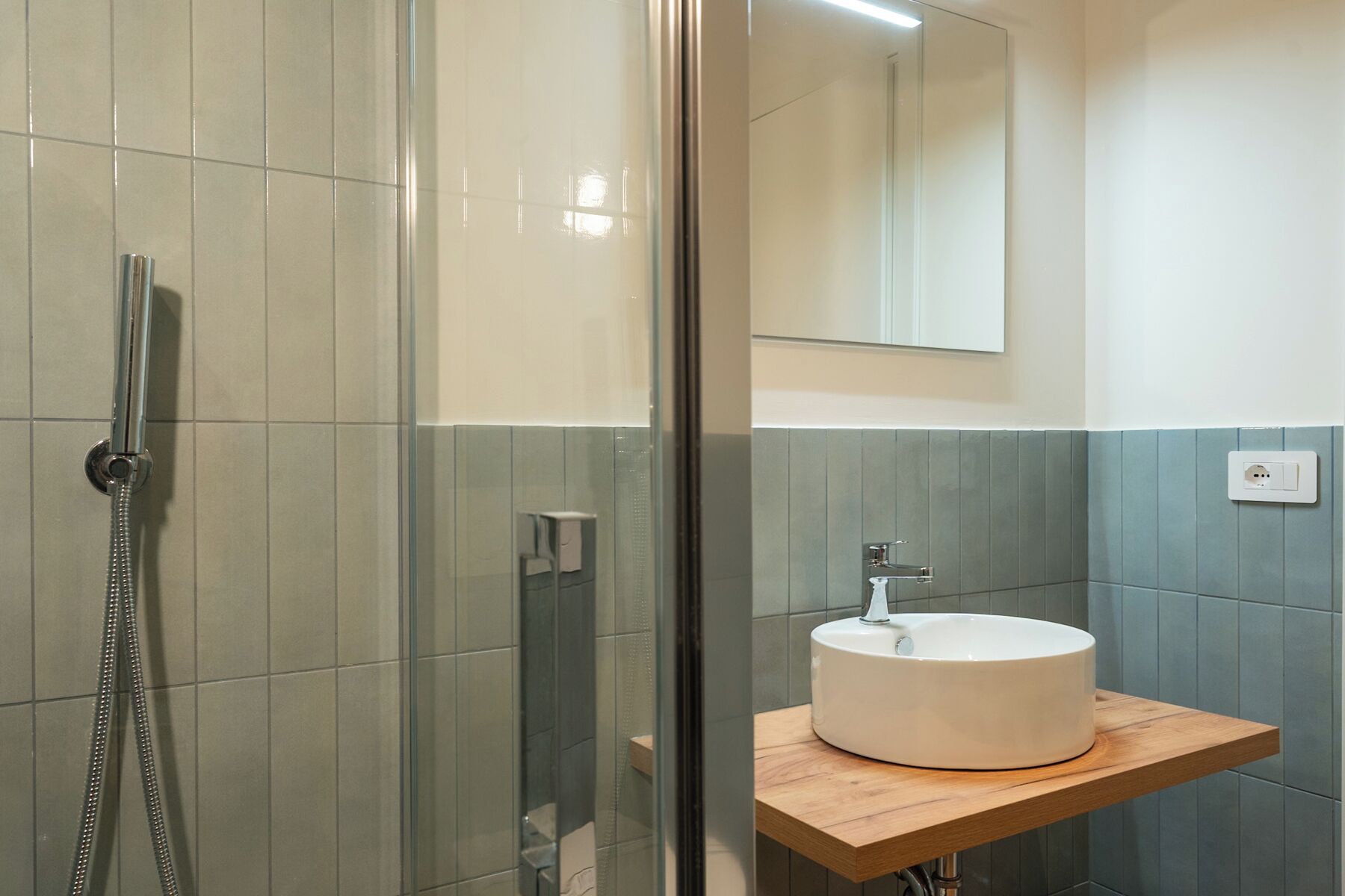 A modern bathroom with a glass-enclosed shower, a white sink on a wooden countertop, and a mirror above the sink.