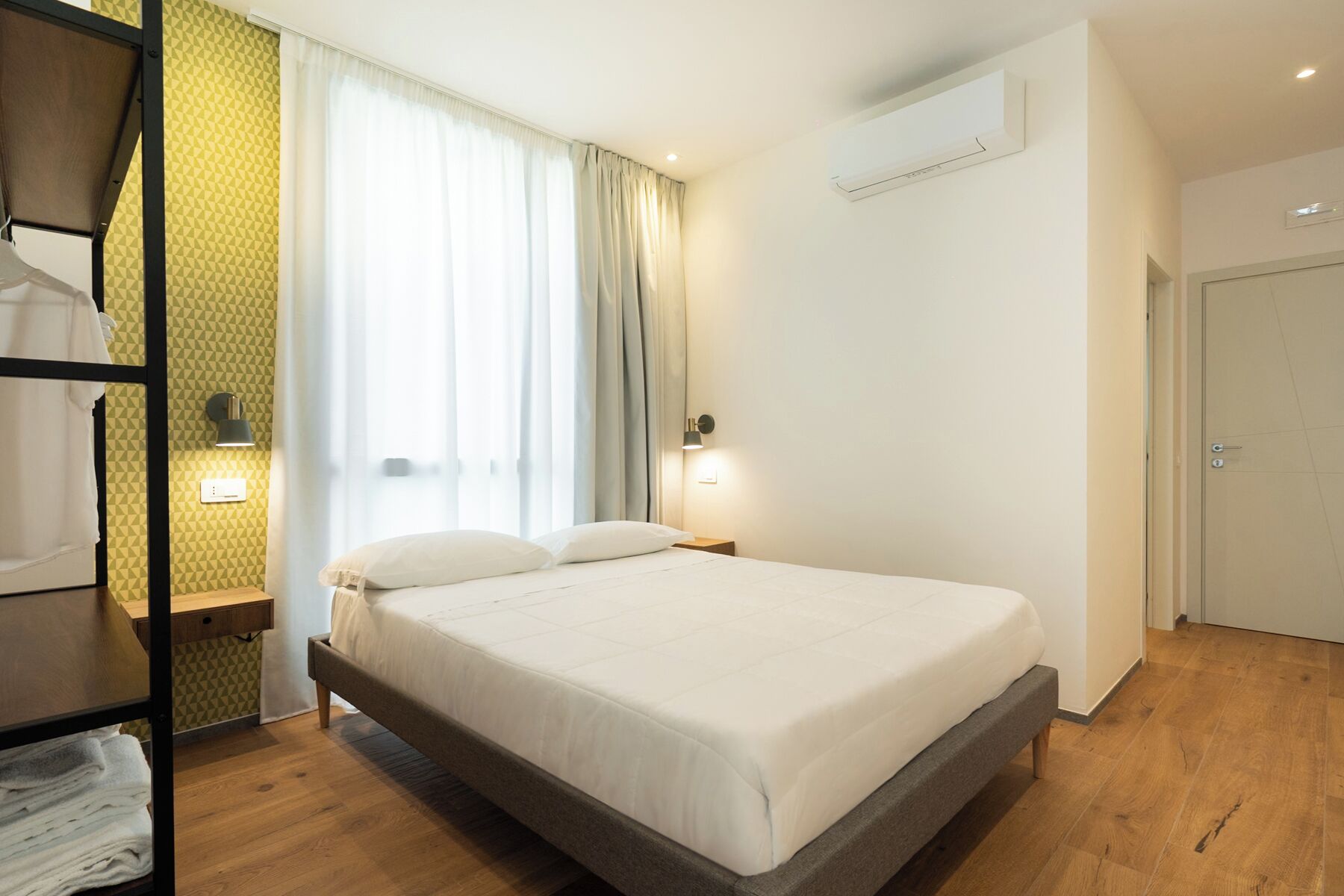 Una cama bien hecha en una habitación de hotel con suelo de madera, una ventana con cortinas, un aire acondicionado de pared y una mesita de noche con lámpara.