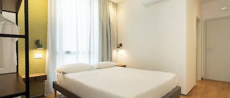 Cama bem arrumada em quarto de hotel, com piso de madeira, janela com cortinas, ar-condicionado fixado na parede e mesa de cabeceira com abajur.