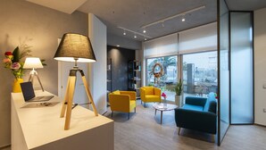 Un moderno vestíbulo de hotel con un mostrador de recepción, una computadora portátil, una lámpara, dos sillones amarillos, un sofá azul y un amplio ventanal con vistas a la ciudad.