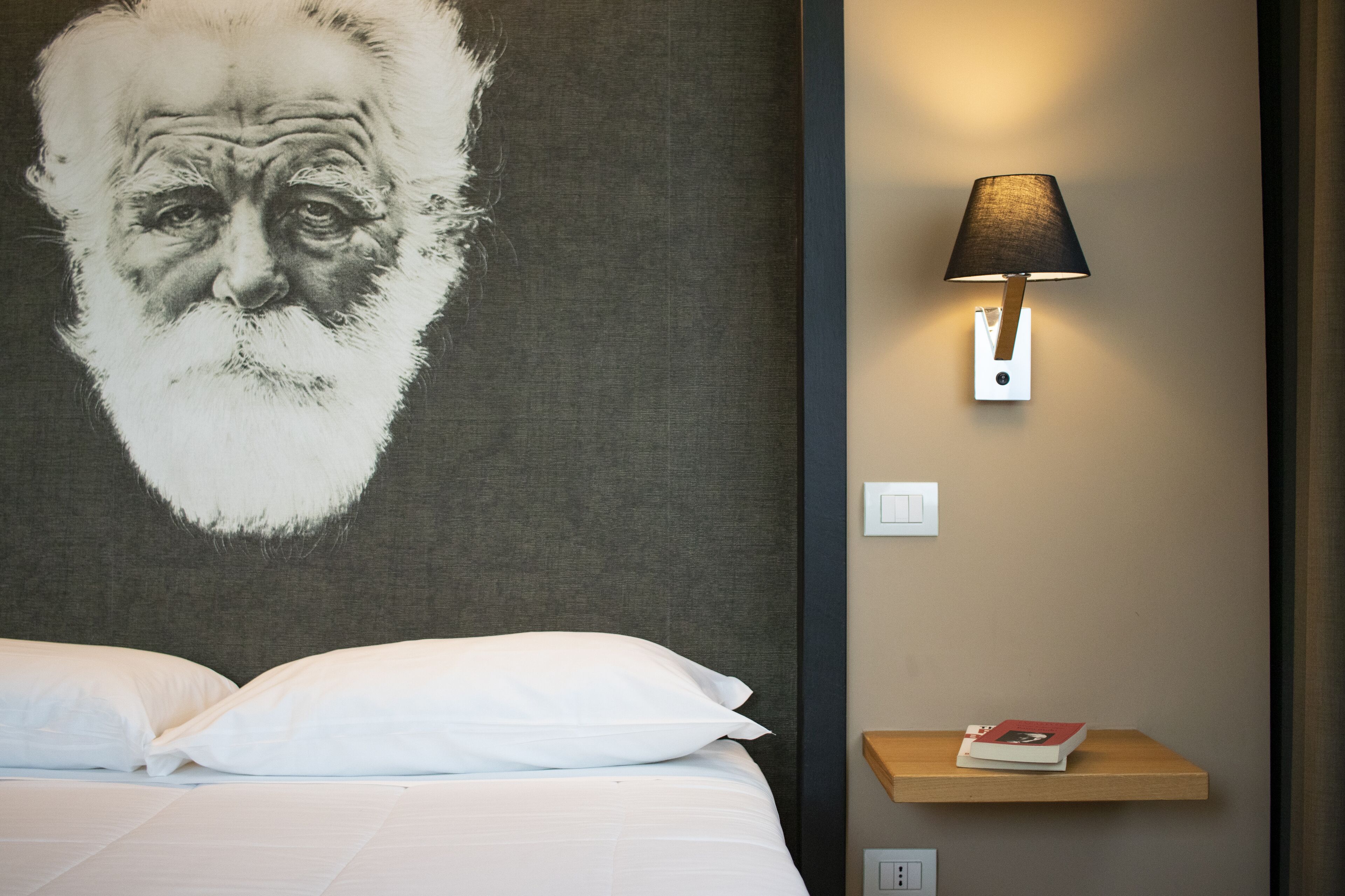 Quarto de hotel com um retrato grande na parede, uma luminária de cabeceira, uma mesa de cabeceira de madeira e uma cama com roupa de cama branca.