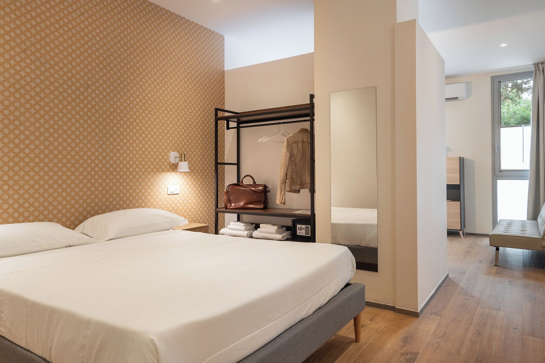 Quarto de hotel moderno com uma cama grande, mesa de cabeceira, guarda-roupa com cabideiro e vista para o exterior pela janela.