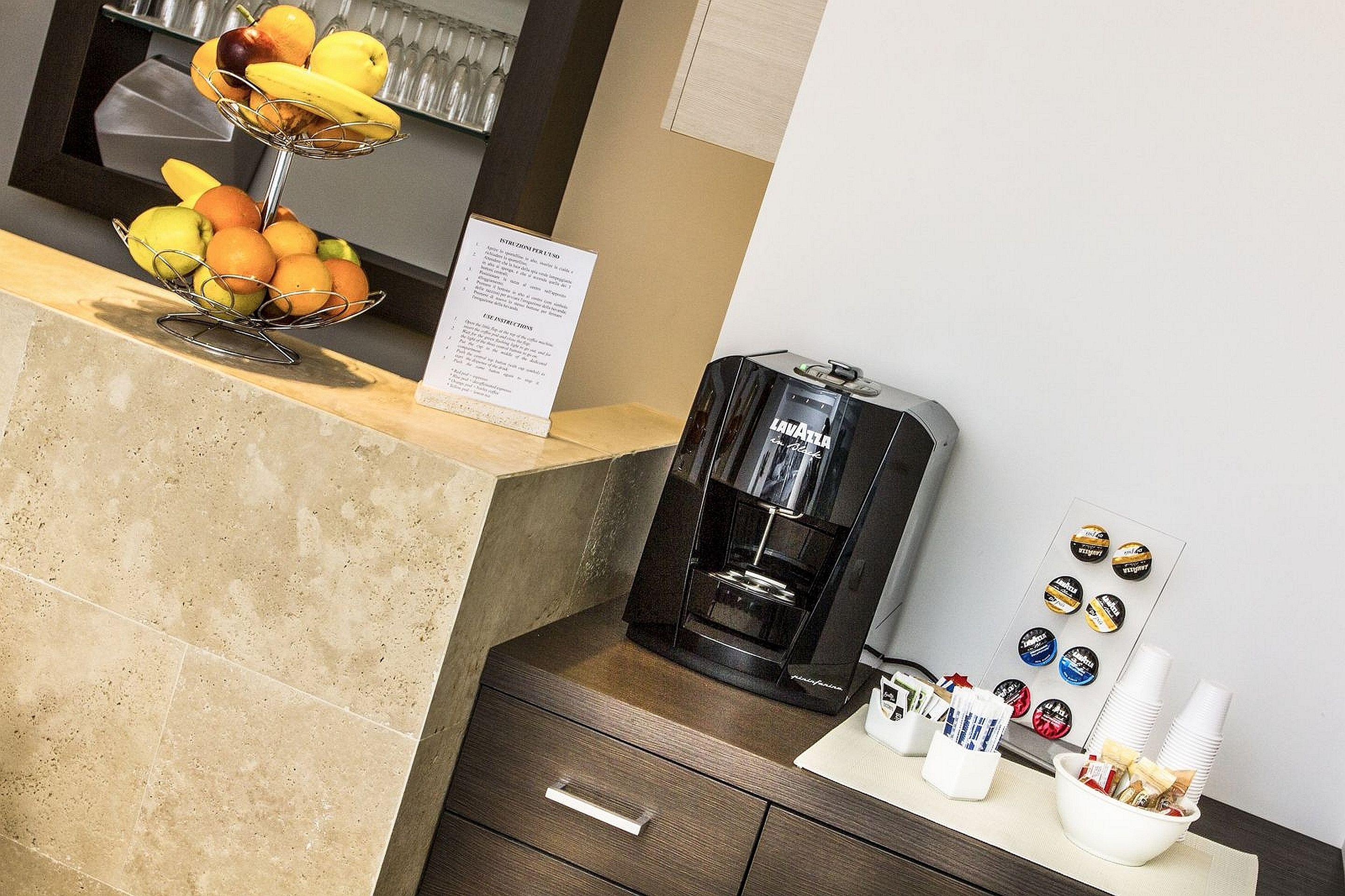Et hotellets reception med et fad med frugt, en kaffemaskine og en opslagstavle med magneter.