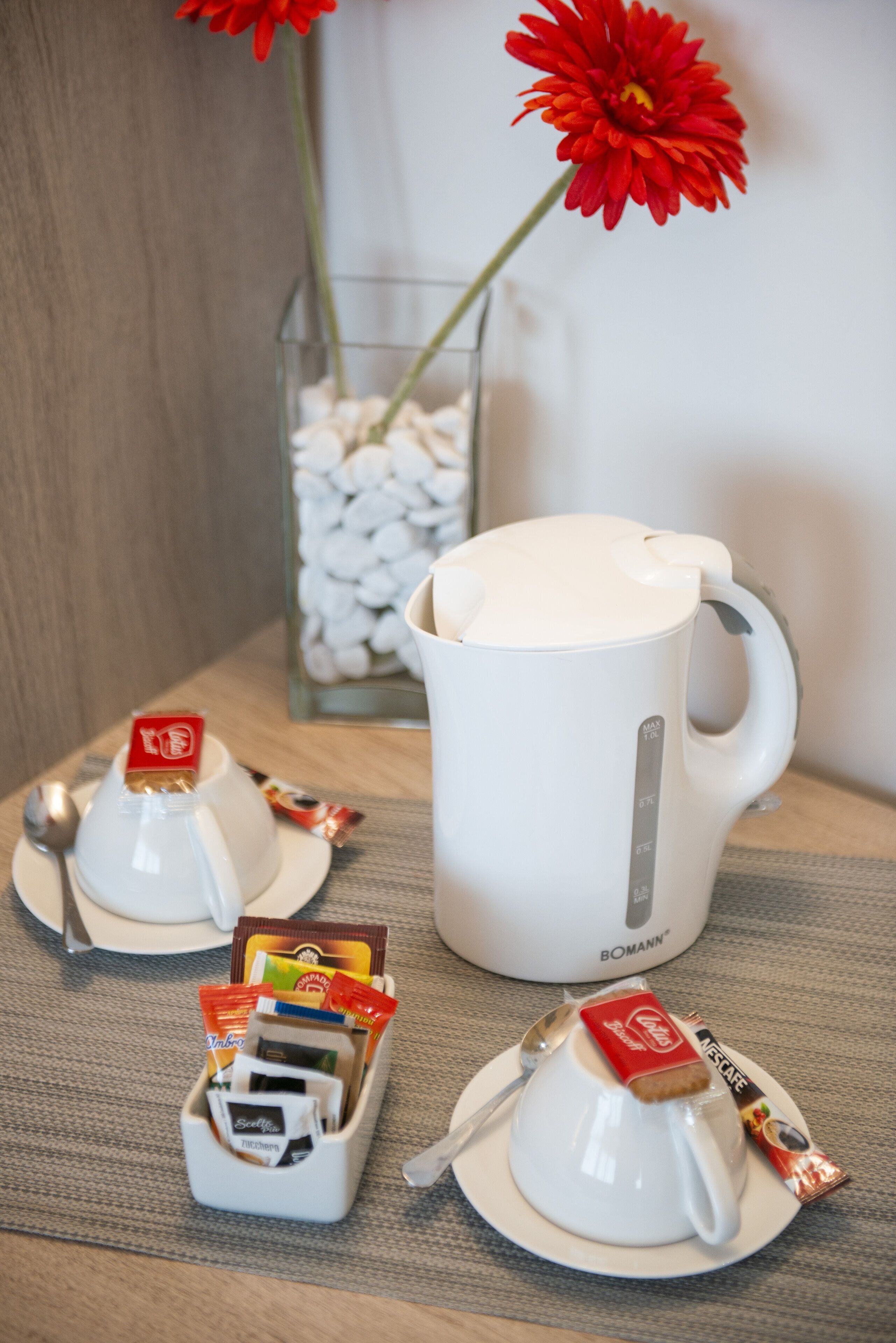 Eine Kaffeebereitstellung mit einer weißen Kaffeemaschine, zwei Tassen und einer Kanne, sowie einem kleinen Behälter mit Kaffeekapseln und einem Löffel.