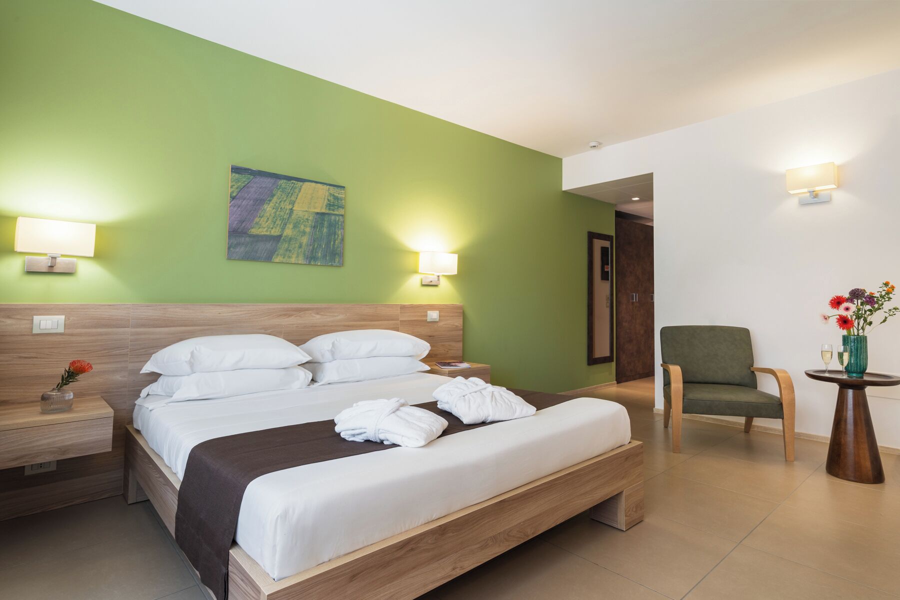 A hotel room with a bed, bedside tables, a chair, a small table with a vase of flowers, and a painting on the wall.