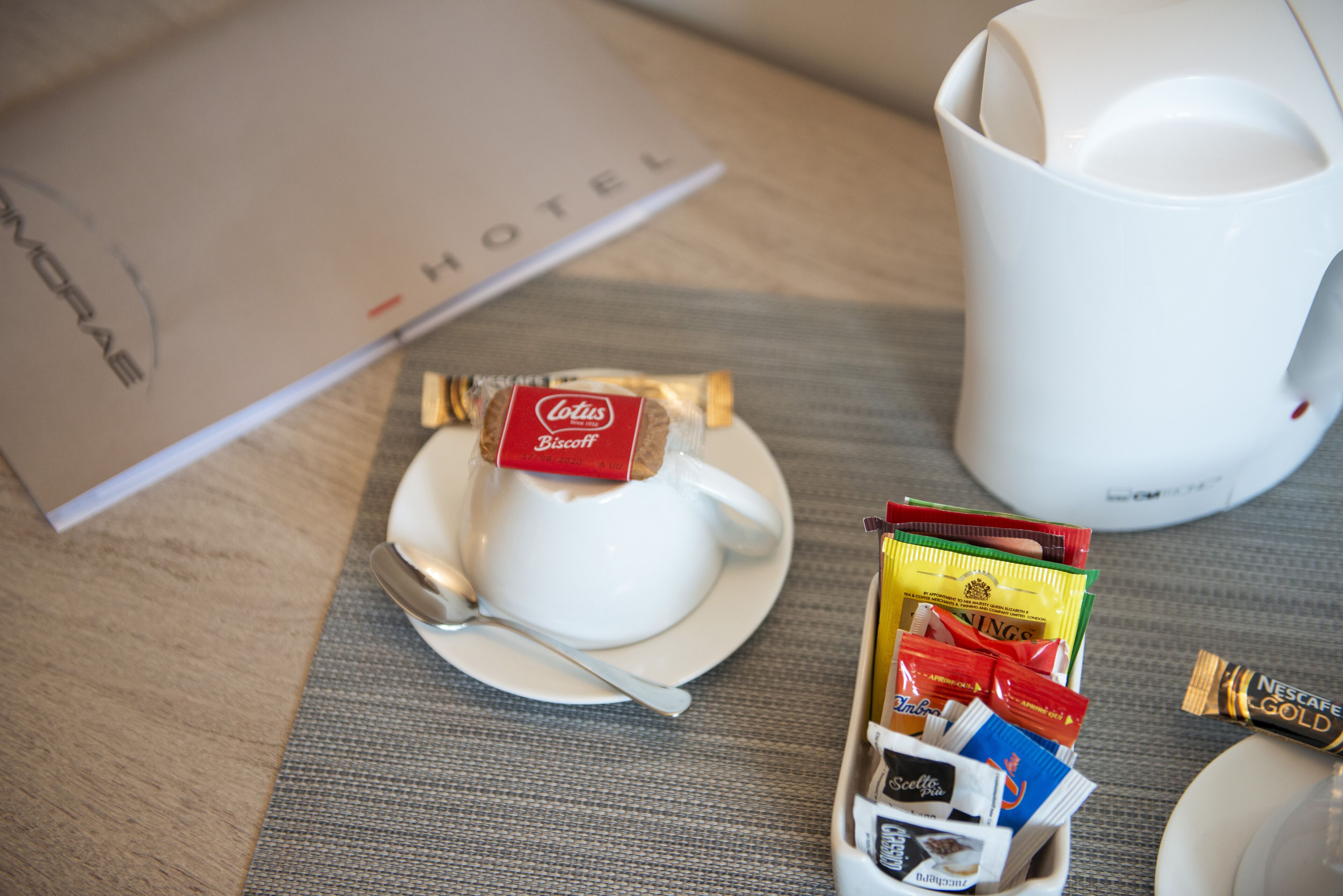 A hotel room with a breakfast setup including a white teapot, a cup, a plate with a spoon, and a tray of assorted snacks and chocolates.