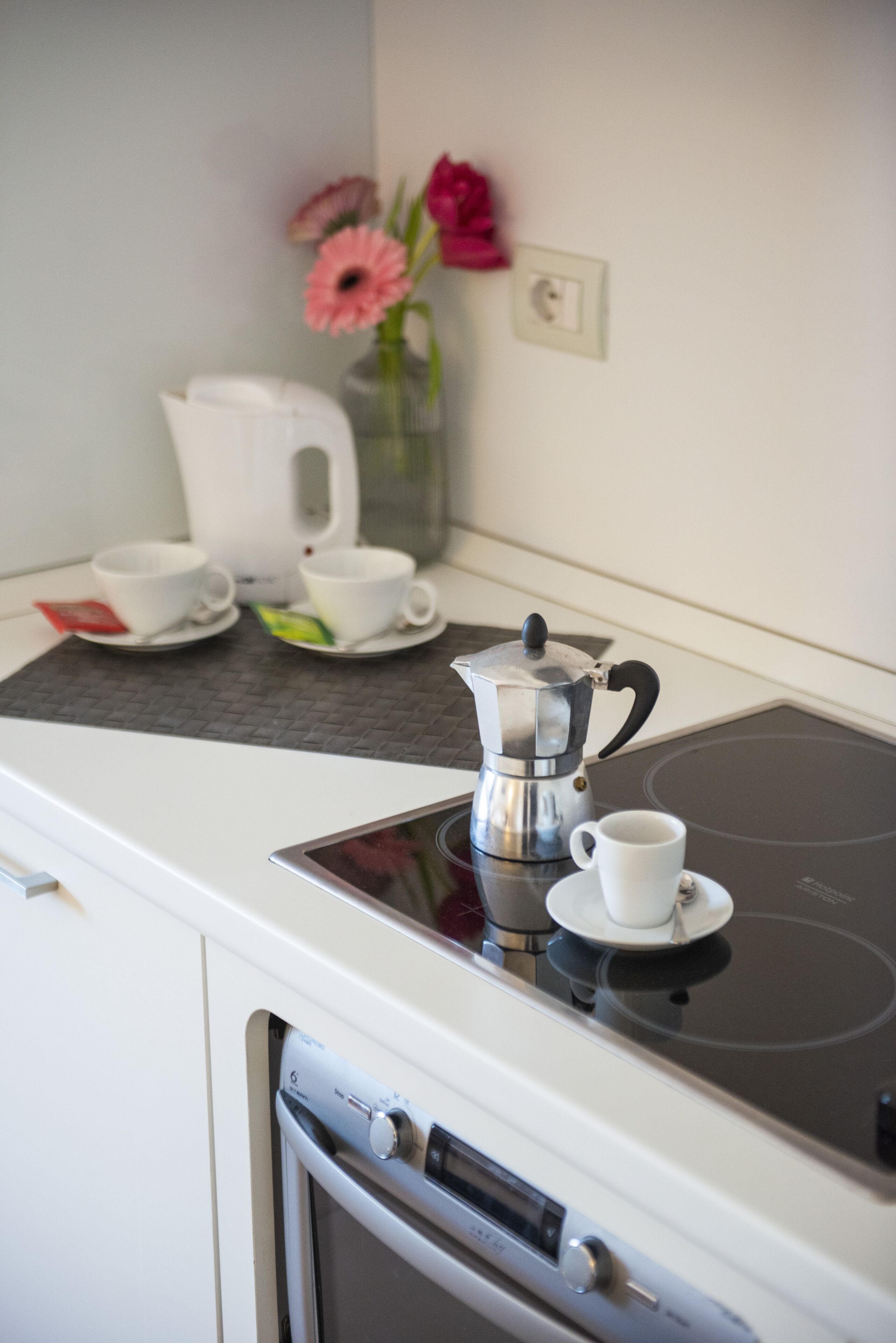 Una cocina con cafetera, dos tazas y un florero con flores sobre la encimera.