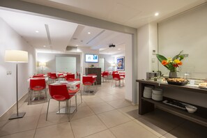 A modern dining area with red chairs and white tables, a buffet counter with food and drinks, and a television screen in the background. - Hotel Dimorae Rooms and Suites - Apartments (Civitanova Marche)