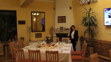 A dining area with a table set for a meal, chairs, a large potted plant, and a television screen displaying a news program.