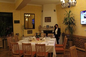 A dining area with a table set for a meal, chairs, a large potted plant, and a television screen displaying a news program.