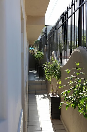 A narrow outdoor corridor with a tiled floor, black metal railing, and potted plants along the walls.