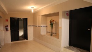 Ein Flur mit Fliesenboden, einem Feuerlöscher, einer Tür und einem an der Wand befestigten Regal mit Blumen.