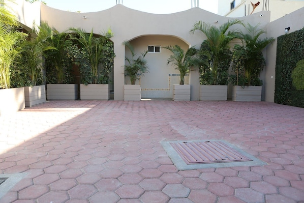A paved courtyard with hexagonal tiles, surrounded by planters with various plants, and a building entrance with a door and window.